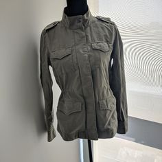 Army (Khaki) Green Utility Jacket From The Gap Dropped Shoulders, Snap-Button Cuffs. Stand Collar, Hidden Front Zip And Snap-Button Closure. Utility Pockets At Chest And Hips Interior Tie-Belt Ties And Cinches At Waist. Stitch Detailing Throughout. "How To Style" Ideas Included Fabric & Care: 97% Cotton, 3% Lycraspandex Machine Wash. Spring Military Outerwear With Stand Collar, Military Style Khaki Utility Jacket With Buttoned Pockets, Khaki Military Outerwear With Button Closure, Khaki Utility Jacket With Snap Buttons For Fall, Military Style Khaki Outerwear With Button Closure, Casual Olive Outerwear With Buttons, Spring Military Outerwear With Buttoned Pockets, Khaki Utility Jacket With Button Cuffs, Military Khaki Outerwear With Buttons