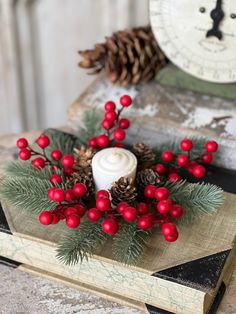 a candle with red berries and pine cones is on top of an old book next to a clock