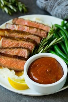 a plate with meat, mashed potatoes and green beans on it next to a small bowl of sauce