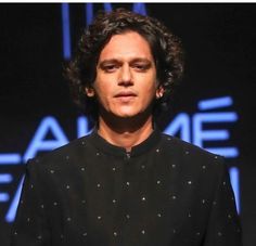 a man with curly hair standing in front of a blue sign and wearing a black shirt