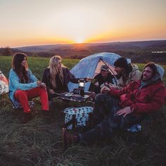 group of people sitting in the grass next to a tent at sunset, with one person holding a cell phone