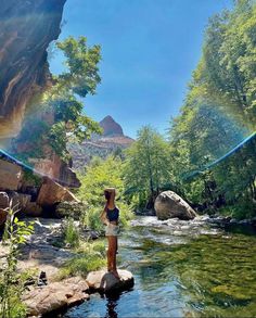 a woman standing on rocks in the middle of a river with mountains in the background