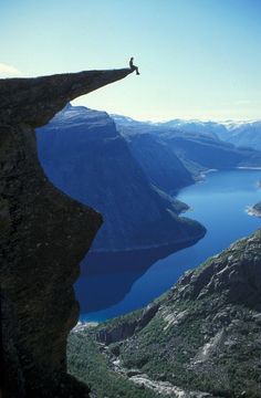 a person standing on top of a cliff next to a body of water with mountains in the background