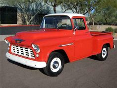 an old red and white truck parked in a parking lot next to a building with trees