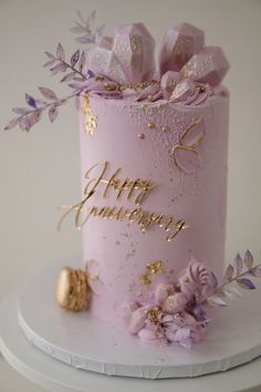 a pink birthday cake with purple flowers and gold lettering on the top, sitting on a white pedestal