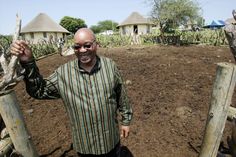 a man is standing in the dirt with his hand up to his face and two thatched huts behind him