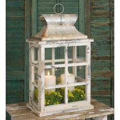 a white lantern with two candles in it on top of a wooden table next to a green wall