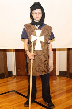 a young boy dressed up in a costume with a cross on it and holding a stick