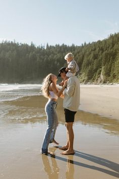 a woman holding a baby while standing on top of a sandy beach next to the ocean