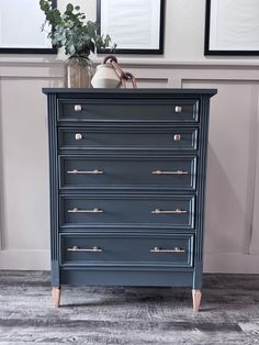 a blue dresser with drawers in front of two framed pictures and a vase on top