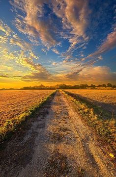 an empty dirt road in the middle of a field with clouds above it at sunset