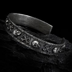 The inspiration for this sterling silver gothic cuff bracelet came from a visit at the Sedlec Ossuary in Czech Republic when my band toured through Europe with Joe Satriani. This small ossuary contains skeleons of betewen 40,000 - 70,000 people artistically arranged into decorations and furnishings. It's truly a stunning macabre location. It features intricate detailing of skulls and bones shaped into a cross encircling the entirety of the bracelet. The frame of the Catacomb Bracelet is designed to resemble the brickwork of the Ossuary's surroundings, adding a touch of realism to this macabre piece. Each skull and femur is expertly crafted to ensure a sense of realism, making the bracelet perfect for those who love to embrace their dark side. I designed this as a matching set to pair with Silver Gothic Skull Bracelets, Silver Punk Cuff Bracelet Gift, Silver Punk Cuff Bracelet For Gift, Silver Punk Bangle Bracelet, Punk Silver Bangle Bracelet, Punk Style Silver Bangle Bracelet, Sedlec Ossuary, Joe Satriani, Bone Ring