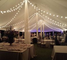 the inside of a tent with tables and chairs covered in white linens is lit by fairy lights