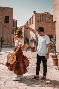 a man standing next to a woman in a brown dress and hat on a cobblestone street