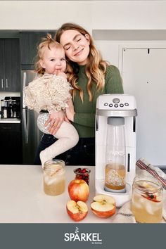 a woman holding a baby in her arms next to an apple juicer and some apples