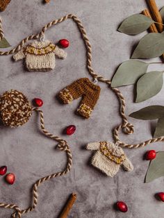 Handmade sweater garland with tan sweaters and brown pants hung on garland with little white wire hangers. Garland string is brown and tan with brown and cream pom poms on each end of the garland. Garland is seen on a concrete counter with cranberries, cinnamon sticks, and greenery. Knit Sweater Ornament, Knitted Garland Christmas, Christmas Tapestry Wall Hangings, Knit Christmas Garland, Diy Crochet Garland, Knitting Christmas Ornaments, Knit Garland, Knitted Garland, Sweater Decorations