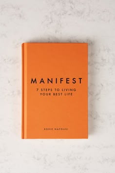 an orange book sitting on top of a white counter next to a marble wall with the title'manfest 7 steps to living your best life '