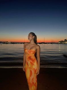 a woman in an orange dress standing on the beach at sunset with boats in the background