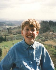 a young boy smiles while standing in front of a field and hills with houses behind him