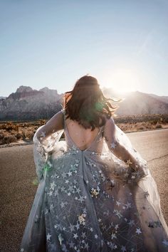 a woman is walking down the road with her dress blowing in the wind and mountains behind her