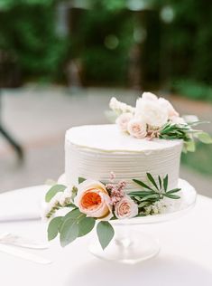 a white wedding cake with flowers on the top is sitting on a table in front of some greenery