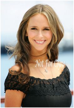 a woman with long hair wearing a black top and smiling at the camera while standing in front of a body of water