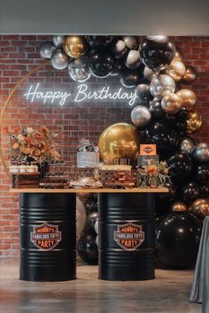 a birthday party with black and gold balloons on the wall behind two large barrel tables