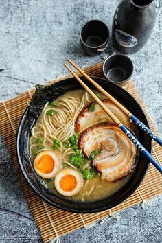 a bowl of ramen with eggs and chopsticks next to two black cups