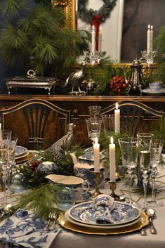 a dining room table set for christmas with blue and white dishes, silver candlesticks and greenery