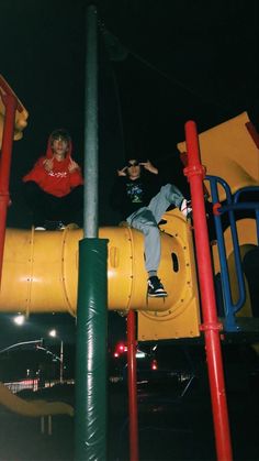 two people sitting on the top of a yellow playground structure at night time with lights shining in the background