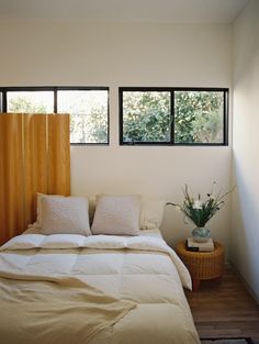 a bed sitting under two windows next to a wooden table with a potted plant on it