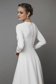 a woman wearing a white dress with pearls on the back and long sleeves, standing in front of a gray background