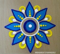 a blue, yellow and white flower on a wooden surface with text that reads meenaka's rangoli