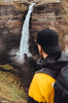 a man standing in front of a waterfall looking at the ground below him and his back to the camera