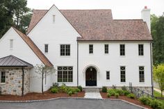 a large white house with a black door and brown shingles on the roof is surrounded by greenery