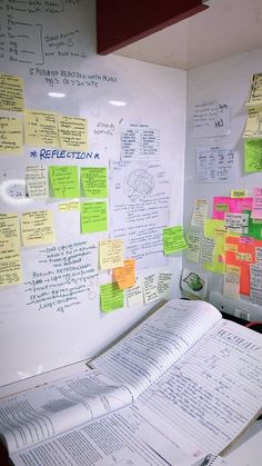 an open book sitting on top of a desk next to a whiteboard covered in sticky notes