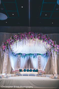 a wedding stage decorated with purple flowers and white draping for the head table