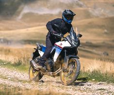 a man riding on the back of a motorcycle down a dirt road next to a field