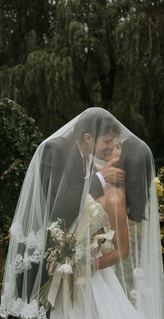 a bride and groom kissing under the veil of their wedding dress in front of trees