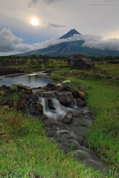 a stream running through a lush green field next to a mountain covered in clouds and grass