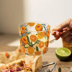 a person is holding a spoon over a glass cup with oranges on it and other food in the background