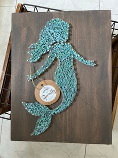 a string art mermaid is hanging on a wooden board with a tag that says,