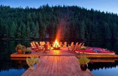 an outdoor fire pit on a dock with chairs around it and trees in the background