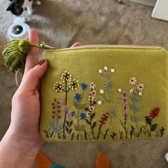 a hand holding a small green purse with flowers on the front and side, while it is in someone's left hand