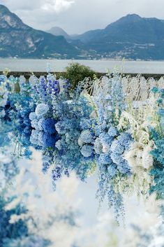 blue and white flowers are lined up on the side of a building near water with mountains in the background