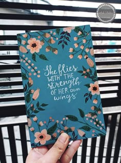 a hand holding up a blue notebook with flowers on it and the words she flies with the strength of her own wings