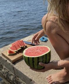 a woman is cutting up a watermelon on the edge of a pier by the ocean