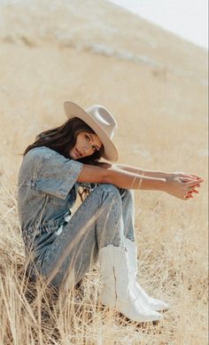 a woman sitting in the middle of a field wearing a cowboy hat and denim overalls