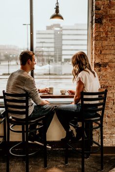 a man and woman sitting at a table in front of a window looking out on the city