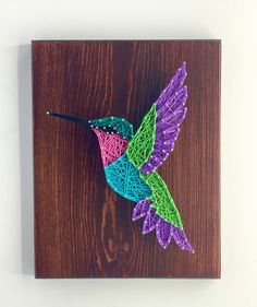 a string art hummingbird sitting on top of a wooden block with purple, green and blue yarn